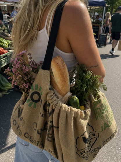 A woman at an outdoor farmers market carries a large burlap grocery tote bag over her shoulder. The tan burlap bag has green printed graphics and text, and is filled with a loaf of bread, fresh herbs, and vegetables.