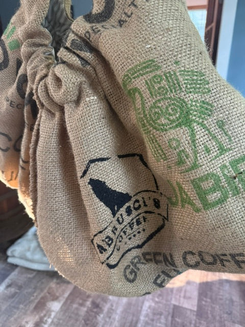 A close-up of a natural burlap drawstring bag printed with green and black coffee-themed graphics and text, including 'ABU SOCI COFFEE' and 'GREEN COFFEE'.