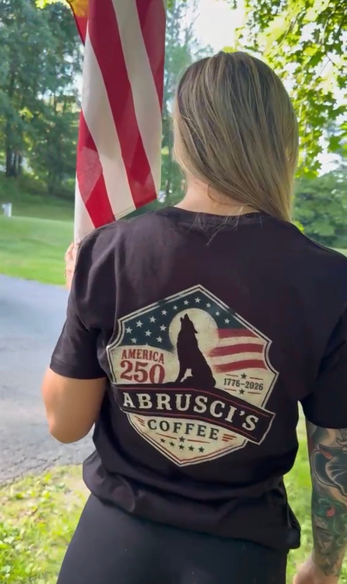 A woman viewed from behind wearing a black t-shirt with a large graphic on the back. The graphic features a wolf silhouette, an American flag, and the text 'ABRUSCI COFFEE' and 'AMERICA 250'.
